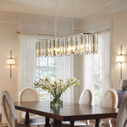 A modern oval-shaped crystal chandelier hanging from a metal frame, with clear, amber, and gray crystal bands, installed in a room with a wooden dining table and chairs.